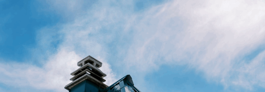 A white cloud of smoke billows from a modern, square chimney on a metal roof against a bright blue sky with scattered clouds.