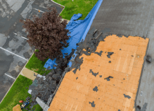 An aerial view shows a damaged roof being repaired, with blue tarps covering some sections and exposed plywood where old shingles have been removed.