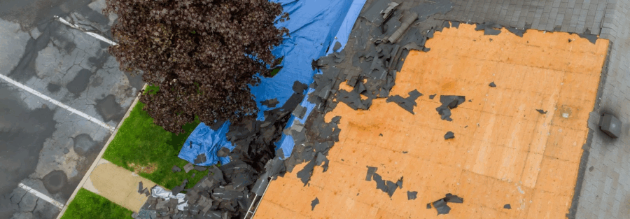 An aerial view shows a damaged roof being repaired, with blue tarps covering some sections and exposed plywood where old shingles have been removed.